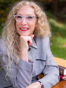 Portrait of Sheree Clark smiling, wearing glasses and a gray blazer, seated at a desk outdoors.